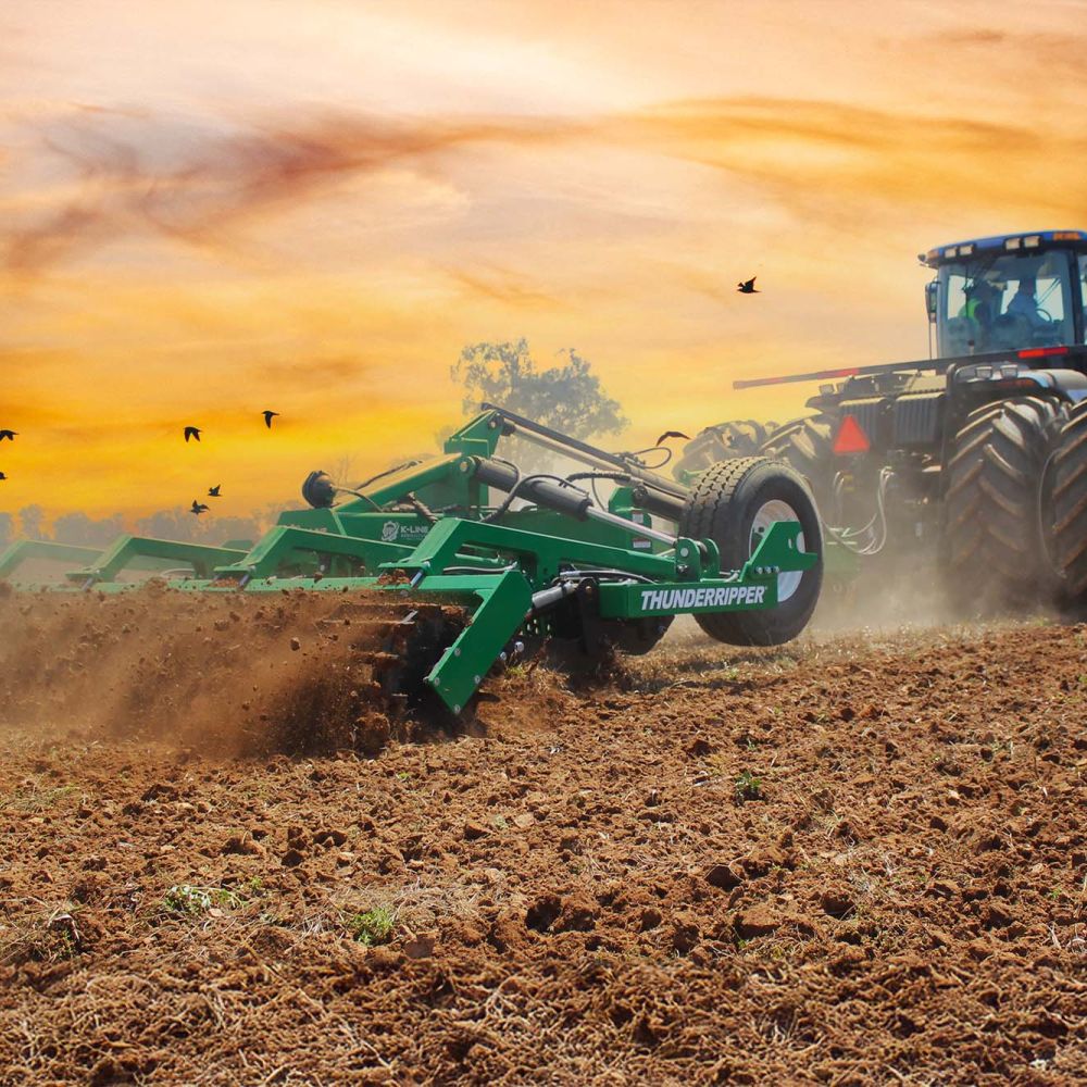 A K-Line Ag ThunderRipper being towed by a New Holland tractor working a large paddock with rick brown soil. There is an intense golden sunset with a flock of birds flying past in the distance.